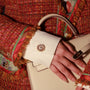 A close-up of a hand resting on a cream-colored leather handbag, featuring Bellis Gold Plated Button Cover on the cuff of a white shirt. The person is wearing a textured red tweed jacket with colorful patterned lining.