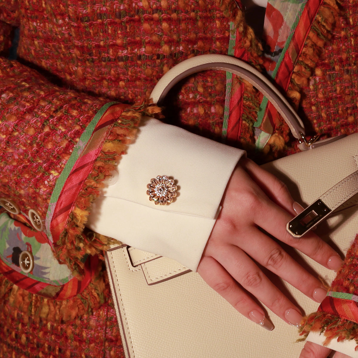 A close-up of a hand resting on a cream-colored leather handbag, featuring Bellis Gold Plated Button Cover on the cuff of a white shirt. The person is wearing a textured red tweed jacket with colorful patterned lining.