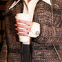 A close-up of a person holding a white paper coffee cup, wearing a grey tweed jacket over a white collared shirt. The jacket has a textured woven pattern and a bellis silver button cover on the cuff.