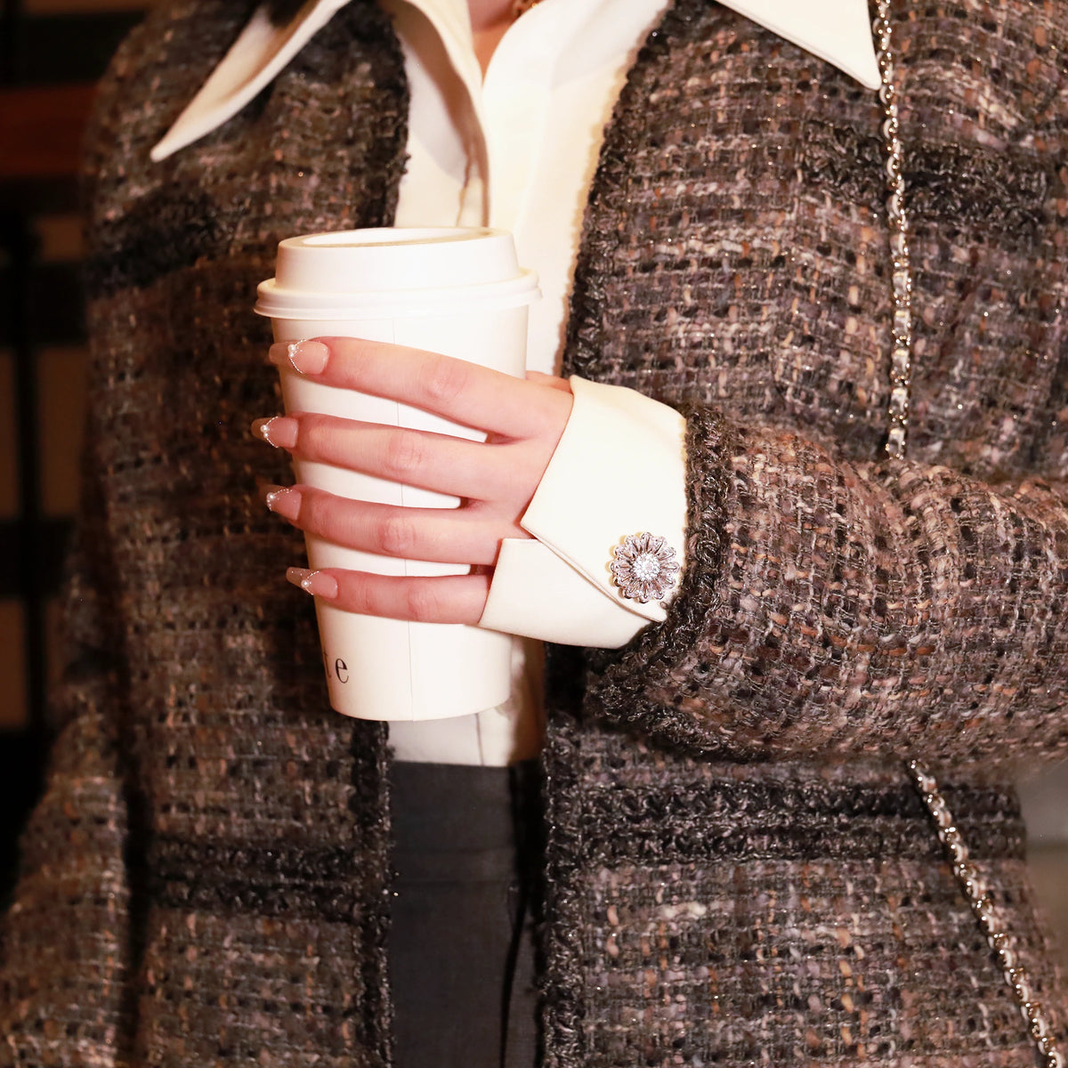 A close-up of a person holding a white paper coffee cup, wearing a grey tweed jacket over a white collared shirt. The jacket has a textured woven pattern and a bellis silver button cover on the cuff.