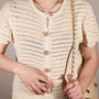 A close-up of a person wearing a cream-colored crochet short-sleeve top with Bellis clip-on button cover from button vena and a keyhole cutout at the collar. The person is also wearing layered gold necklaces and holding a gold chain strap.