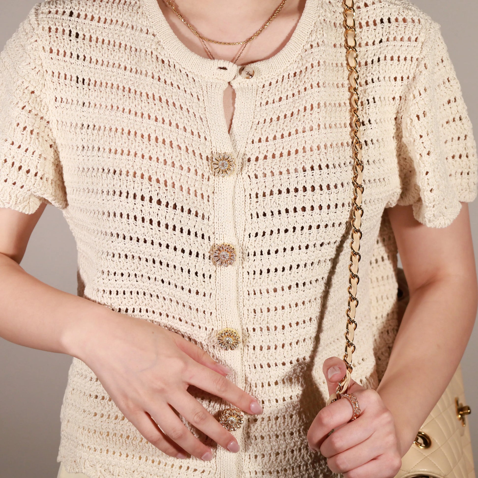 A close-up of a person wearing a cream-colored crochet short-sleeve top with Bellis clip-on button cover from button vena and a keyhole cutout at the collar. The person is also wearing layered gold necklaces and holding a gold chain strap.