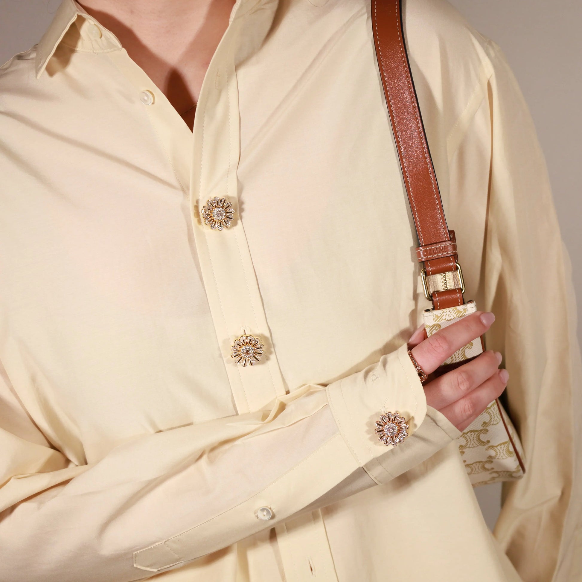 A close-up of a person wearing a cream-colored long-sleeve button-up shirt with decorative Bellis clip-on button cover and holding a brown leather shoulder bag strap.