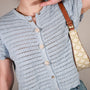 A close-up of a person wearing a light blue crocheted short-sleeve button-front top with textured buttons, paired with a patterned shoulder bag and a brown leather strap, against a plain beige background.