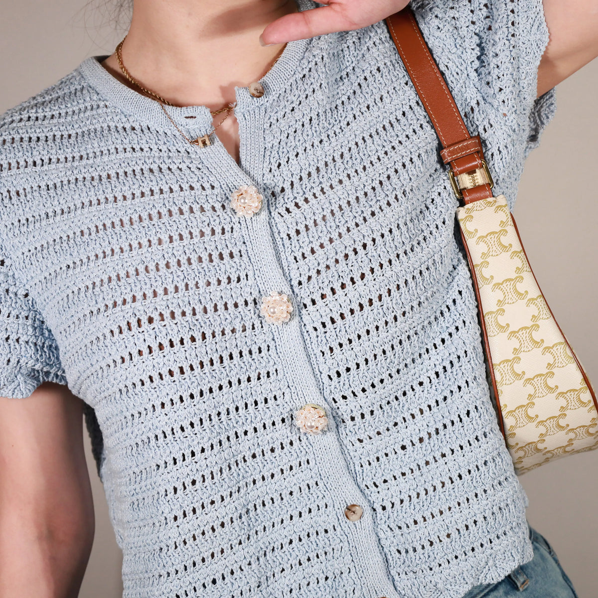 A close-up of a person wearing a light blue crocheted short-sleeve button-front top with textured buttons, paired with a patterned shoulder bag and a brown leather strap, against a plain beige background.
