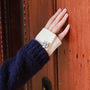 A close-up of a person's hand resting on a warm brown wooden door, wearing a  sweater layer with a silver glacier blossom silver plated button cover and a white shirt