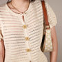 A close-up of a person wearing a cream-colored crochet knit top with Retro Dahlia clip-on button covers, carrying a small cream shoulder bag with a gold monogram pattern and brown leather trim.
