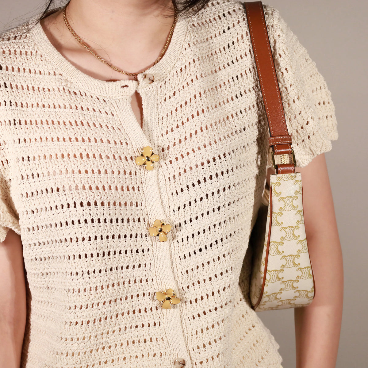 A close-up of a person wearing a cream-colored crochet knit top with Retro Dahlia clip-on button covers, carrying a small cream shoulder bag with a gold monogram pattern and brown leather trim.