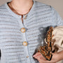 A close-up of a person wearing a light blue open-knit cardigan with riviera pearl button covers from button vena, holding a beige bag with a chunky gold chain strap.