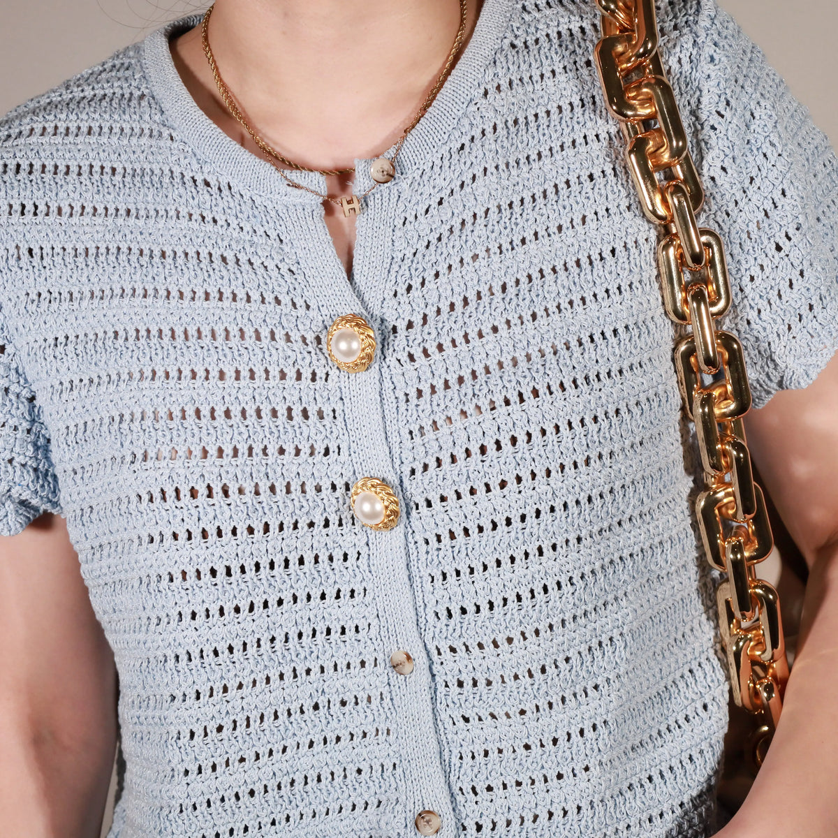 Close-up of a person wearing a light blue open-knit short-sleeve cardigan with riviera pearl button cover from button vena, a gold chain necklace, and carrying a brown shoulder bag with a chunky gold chain strap.