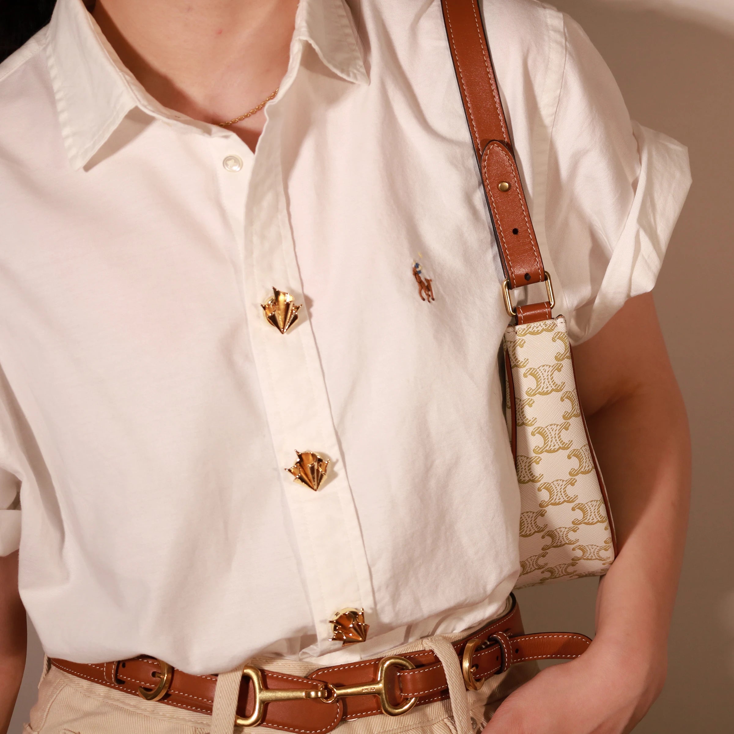 A close-up lifestyle shot of a person wearing a white short-sleeve button-up shirt with decorative gold-tone foulard buttons covers, a brown leather belt, and a small beige crossbody bag with a brown leather strap.