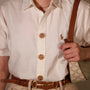 A close-up of a person wearing a cream-colored short-sleeve button-up shirt with decorative gold-toned midnight spark button covers and a small embroidered polo player logo on the chest. The person is also wearing a brown leather belt and holding a patterned shoulder bag.
