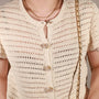 Close-up of a person wearing a cream open-knit short-sleeve cardigan with pearl constellation button cover and layered gold necklaces, against a neutral light gray background.