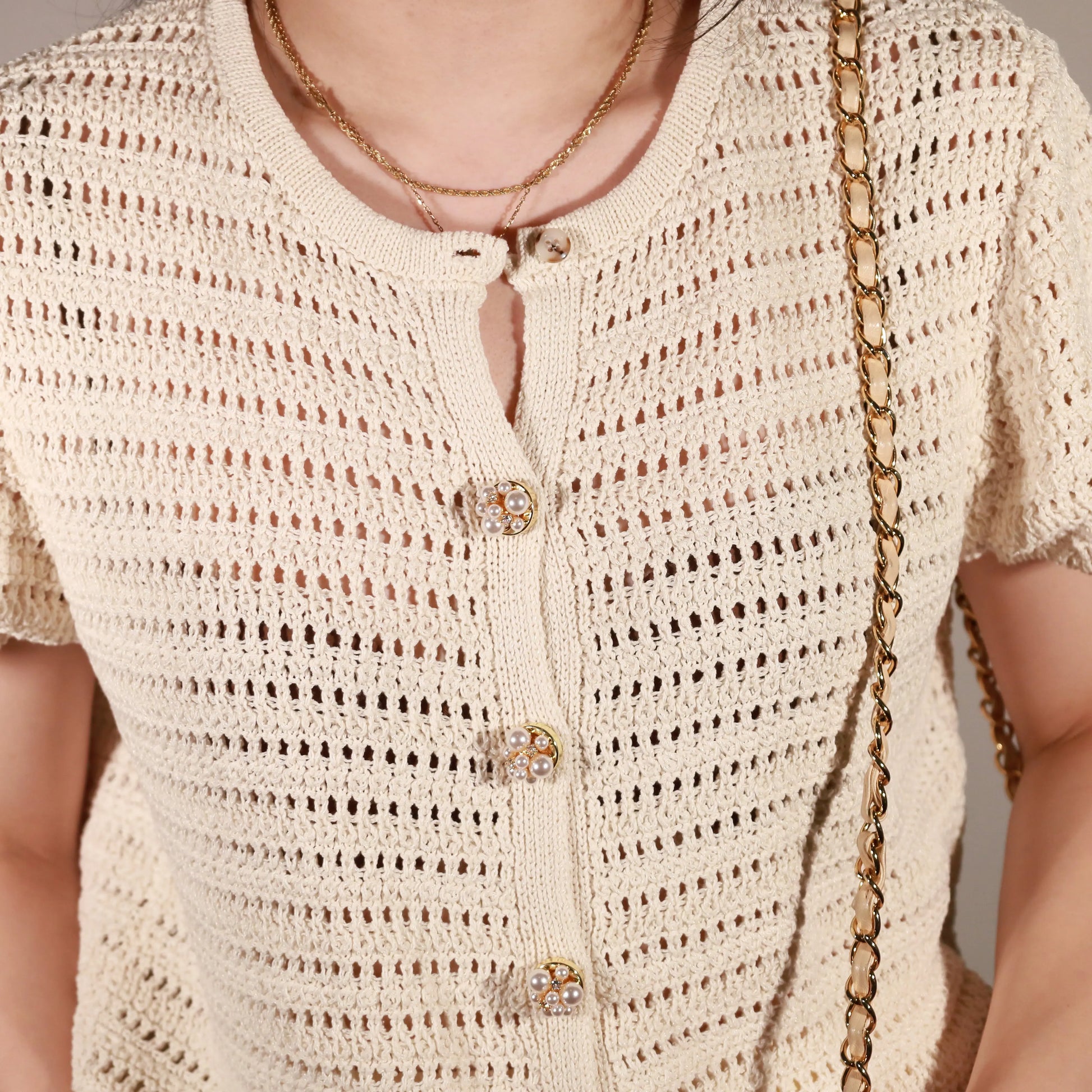 Close-up of a person wearing a cream open-knit short-sleeve cardigan with pearl constellation button cover and layered gold necklaces, against a neutral light gray background.