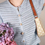 A close-up of a person wearing a light blue open-knit crochet cardigan with decorative pearl constellation button cover, carrying a cream shoulder bag with a gold monogram pattern and brown leather strap. Purple flowers are held in one hand against a soft beige background.